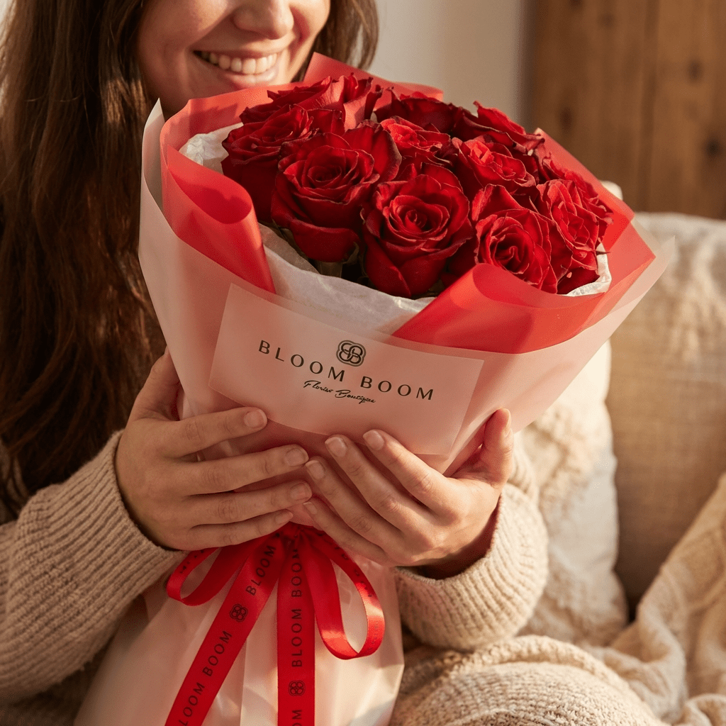 Crimson Promise flower arrangement - a lush bouquet of deep red roses wrapped in translucent branded paper and tied with a red ribbon held by a smiling woman