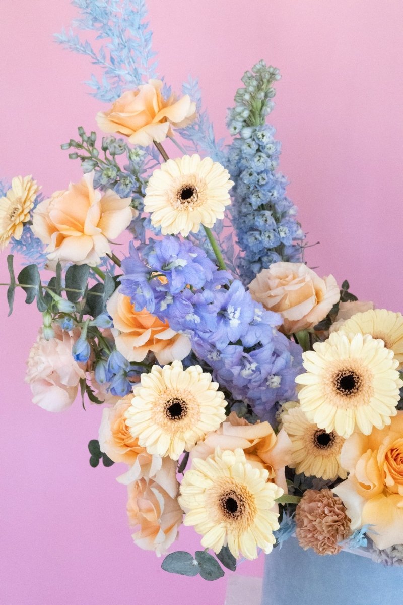 Azure dream flower box with peach roses, cream gerbera daisies, blue delphiniums and eucalyptus on pink background