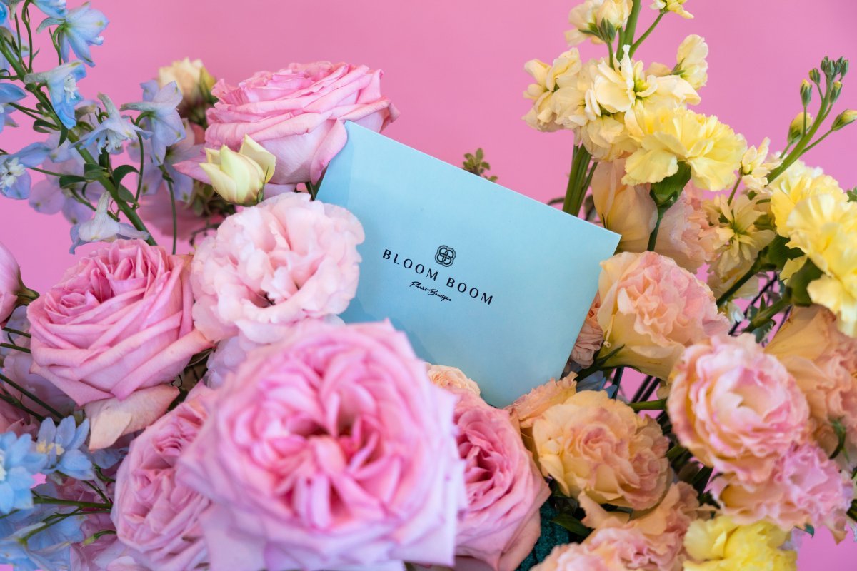 Blue Bloom Boom gift card surrounded by pink roses, yellow carnations, and blue delphinium on pink background