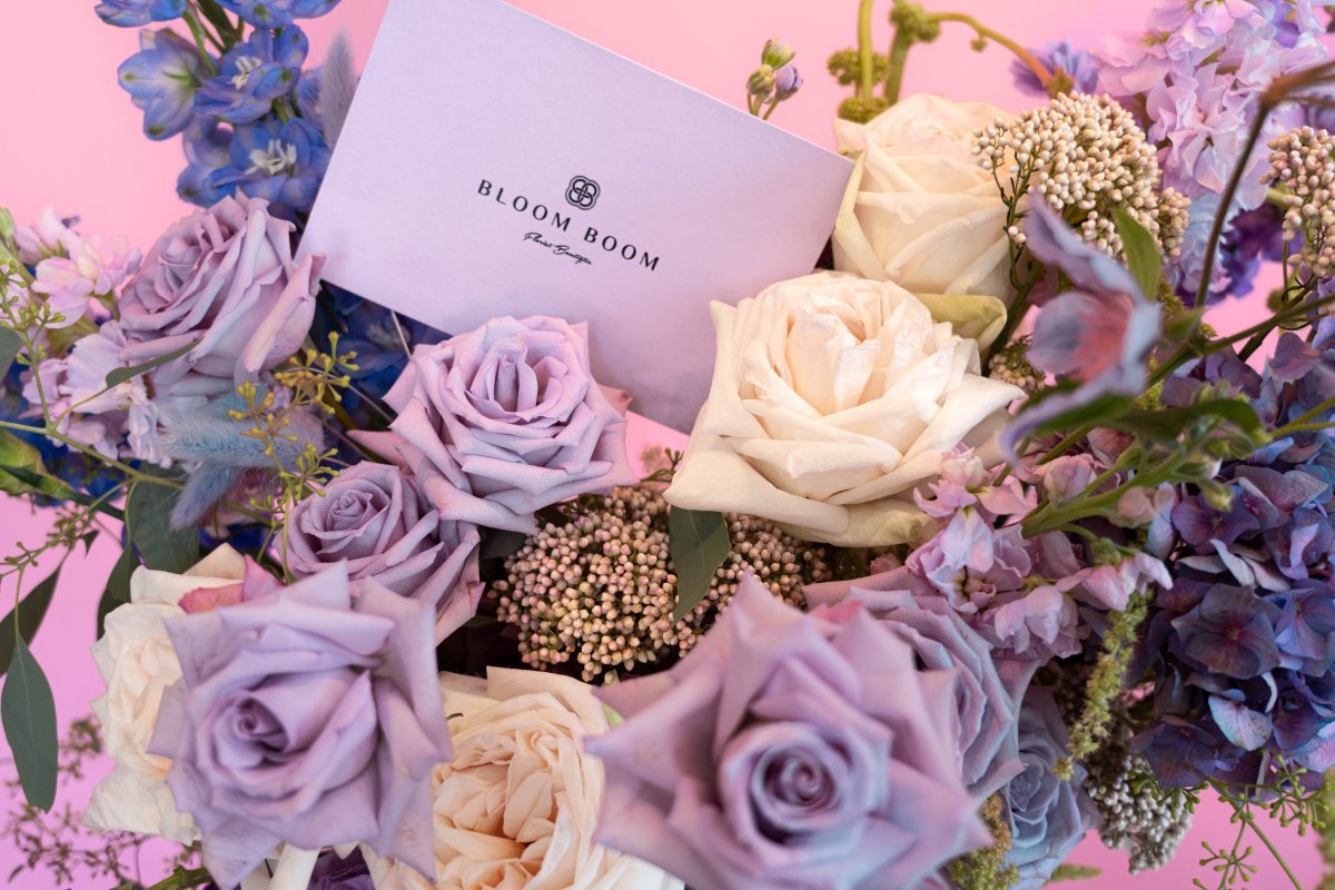Bloom Boom luxury bouquet with lavender roses, cream roses, blue delphiniums and baby's breath on pink background
