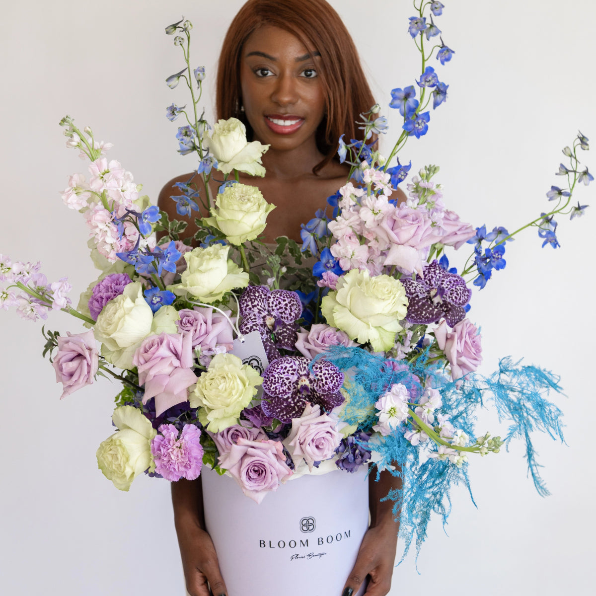 Large white Bloom Boom box filled with purple roses, cream roses, blue delphiniums, and turquoise dried grasses