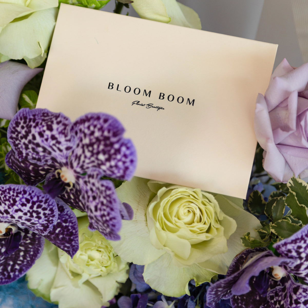 Bloom Boom flower bouquet with purple spotted orchids, cream roses, and pink ribbon on branded card