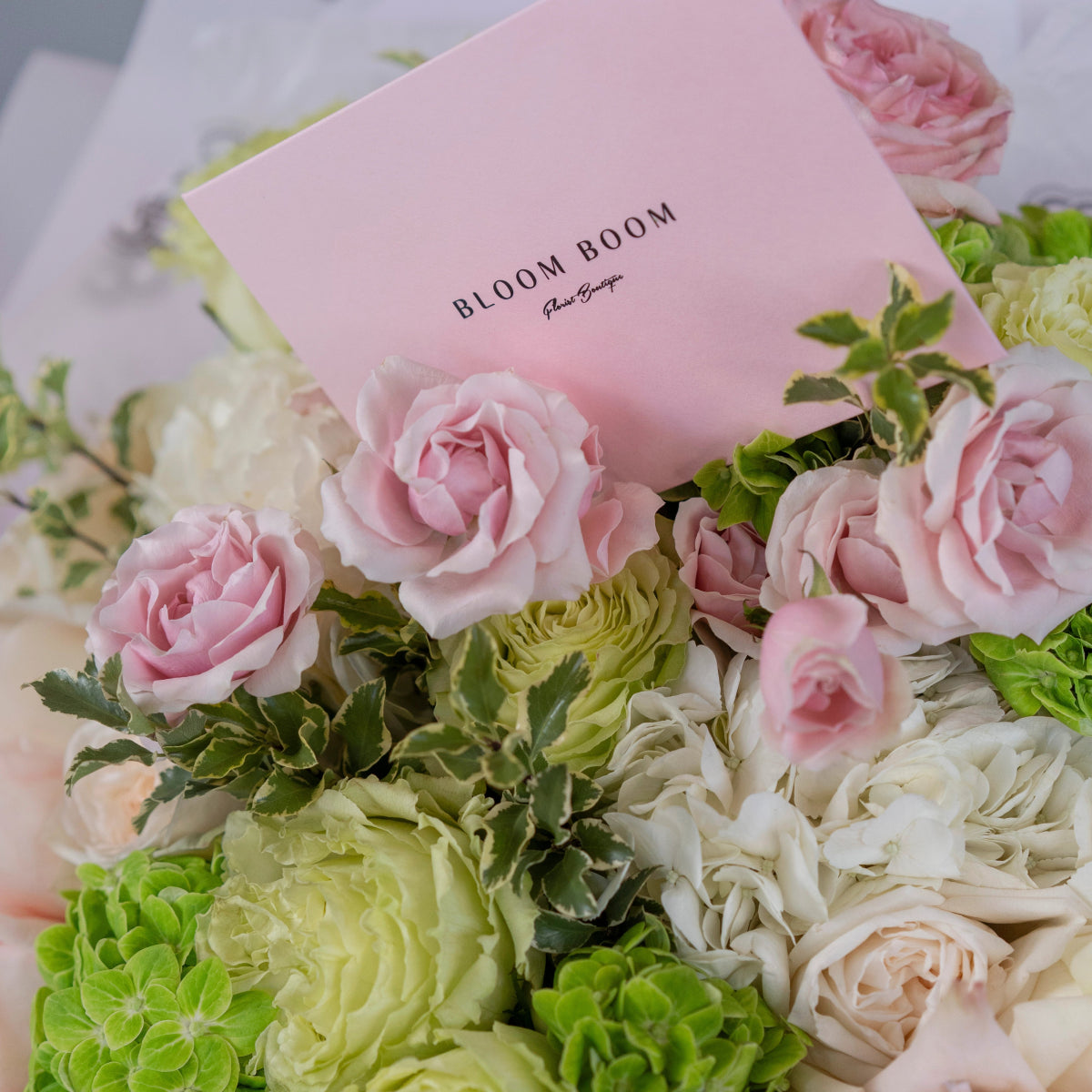 Pink roses, white ranunculus and green hydrangea bouquet with Bloom Boom pink gift card on white marble surface