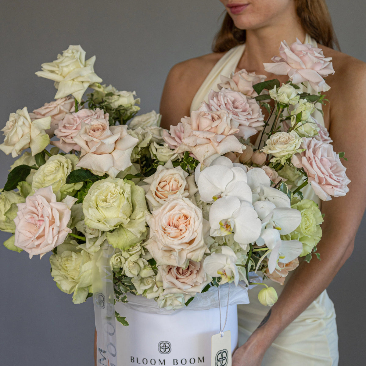 Luxury white flower box with blush pink roses, white orchids, and green hydrangeas held by woman in cream dress