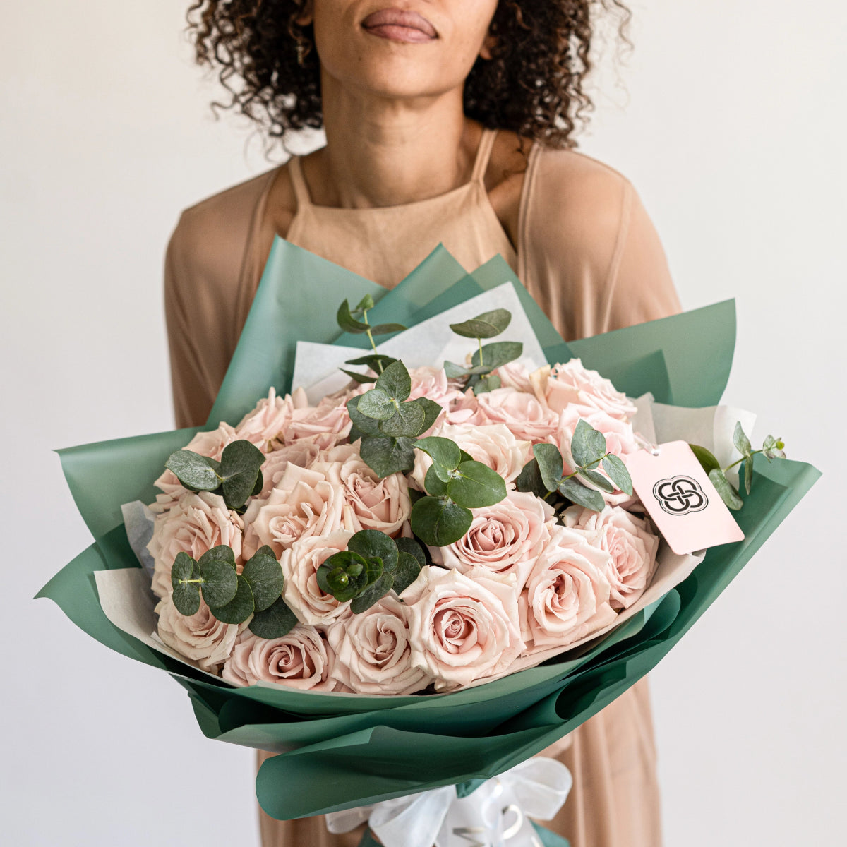 Blush pink rose bouquet with eucalyptus leaves wrapped in sage green paper, held by woman in beige dress