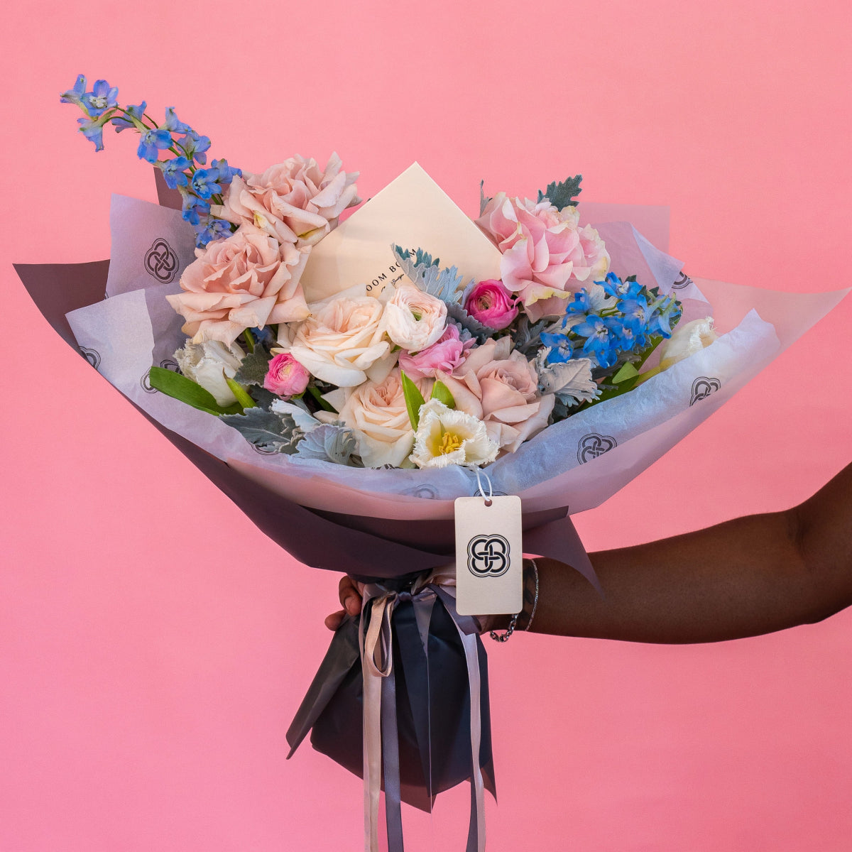 Mixed flower bouquet with pink roses, blue delphinium, white flowers wrapped in purple paper with black ribbon