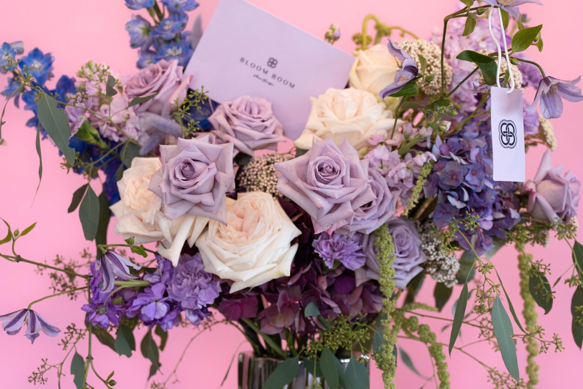 Elegant bouquet with lavender and cream roses, blue delphiniums, purple accents in clear glass vase on pink backdrop
