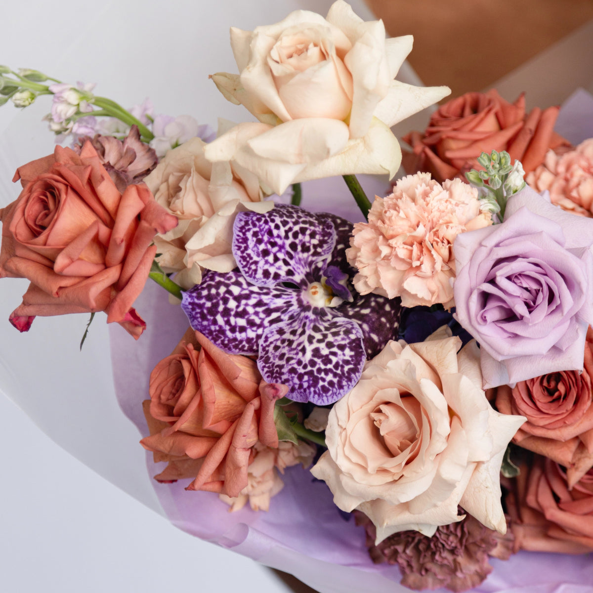 Mixed flower bouquet with coral roses, lavender roses, purple spotted orchids, pink carnations in lavender wrapping