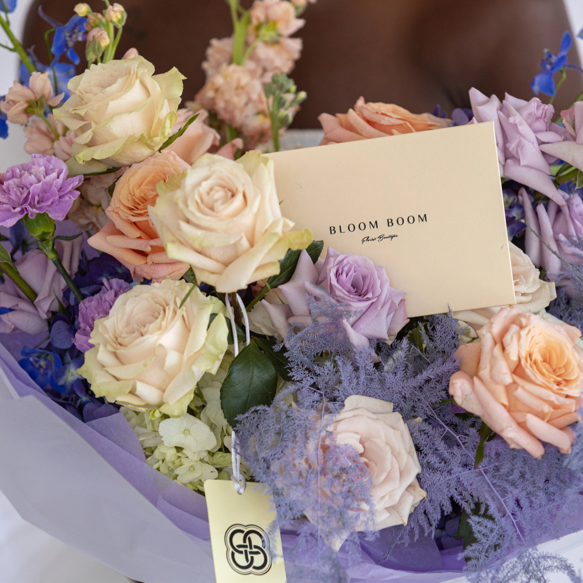 Pastel mixed flower bouquet with peach roses, purple blooms, dusty miller in lavender wrapping with Bloom Boom card