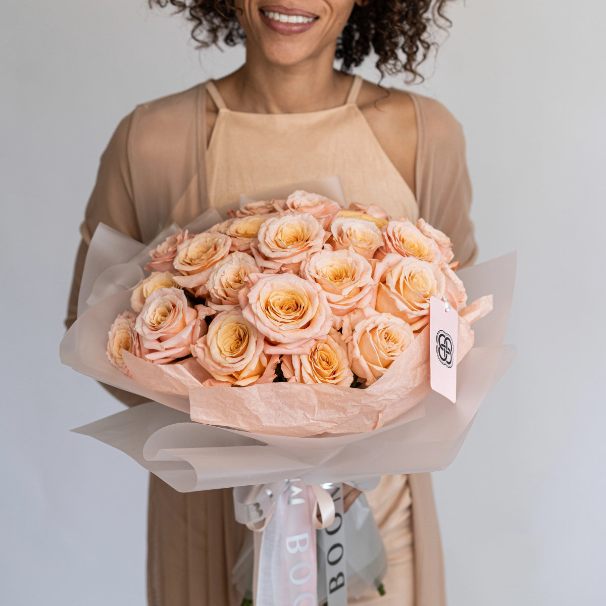 Elegant peach and coral rose bouquet wrapped in translucent paper with ribbon, held by smiling woman in beige outfit