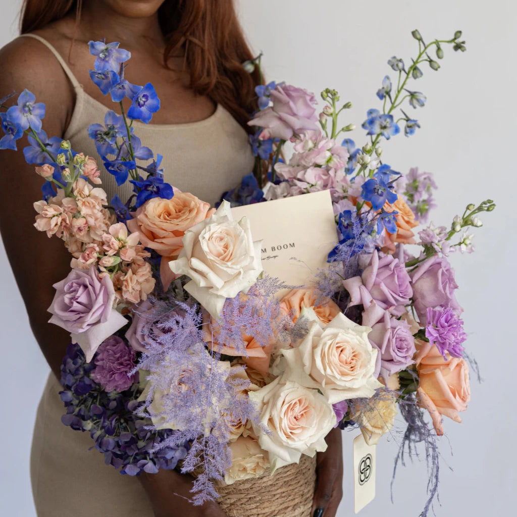 Rustic wicker basket filled with peach roses, purple hydrangea, blue delphinium and lavender flowers with ribbon