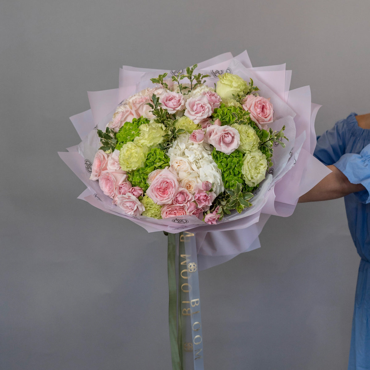 Pink roses and white hydrangea bouquet wrapped in lavender paper with green foliage and decorative ribbon