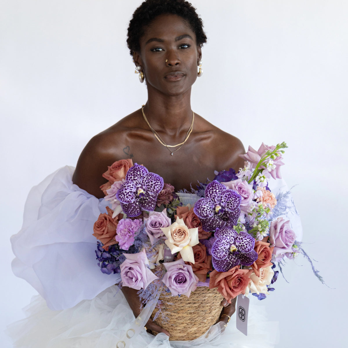 Wicker basket arrangement with purple spotted orchids, lavender roses, coral roses and white tissue paper wrapping