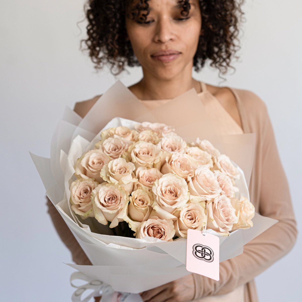 Large bouquet of blush pink quicksand roses wrapped in white paper with brand tag held by woman in beige top
