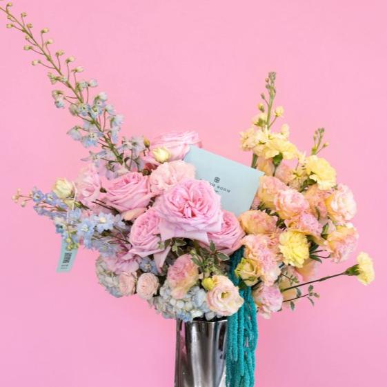 Mixed spring bouquet with pink roses, yellow carnations, blue delphinium in chrome vase against pink backdrop