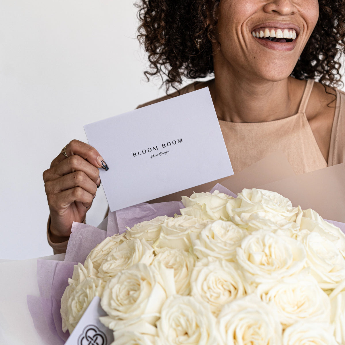 Smiling woman in beige top holding white roses bouquet with Bloom Boom branded card in lavender tissue paper