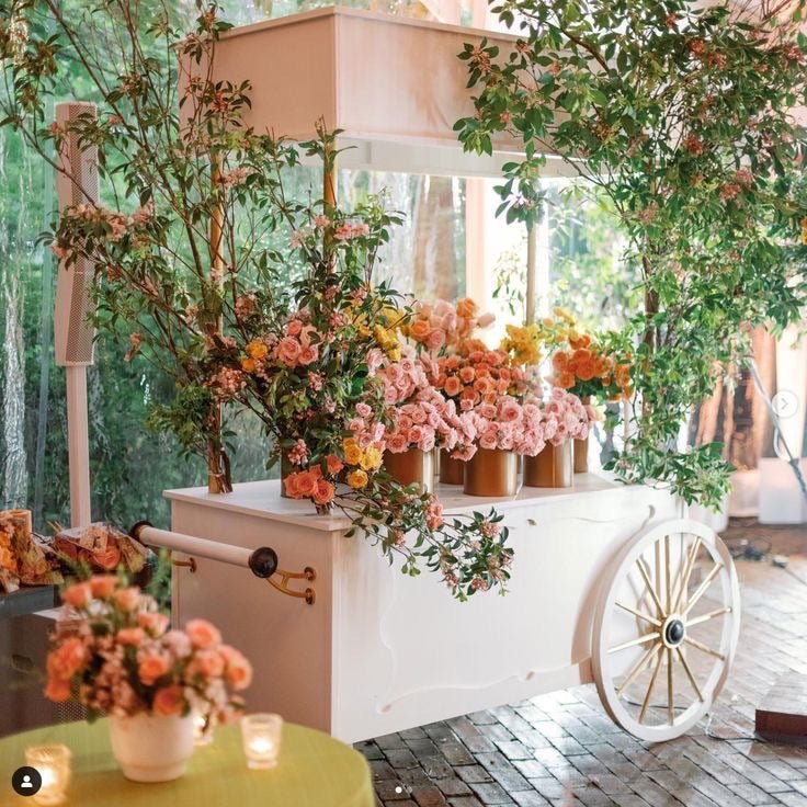 White wooden flower cart with pink coral orange blooms in copper buckets surrounded by greenery in greenhouse setting