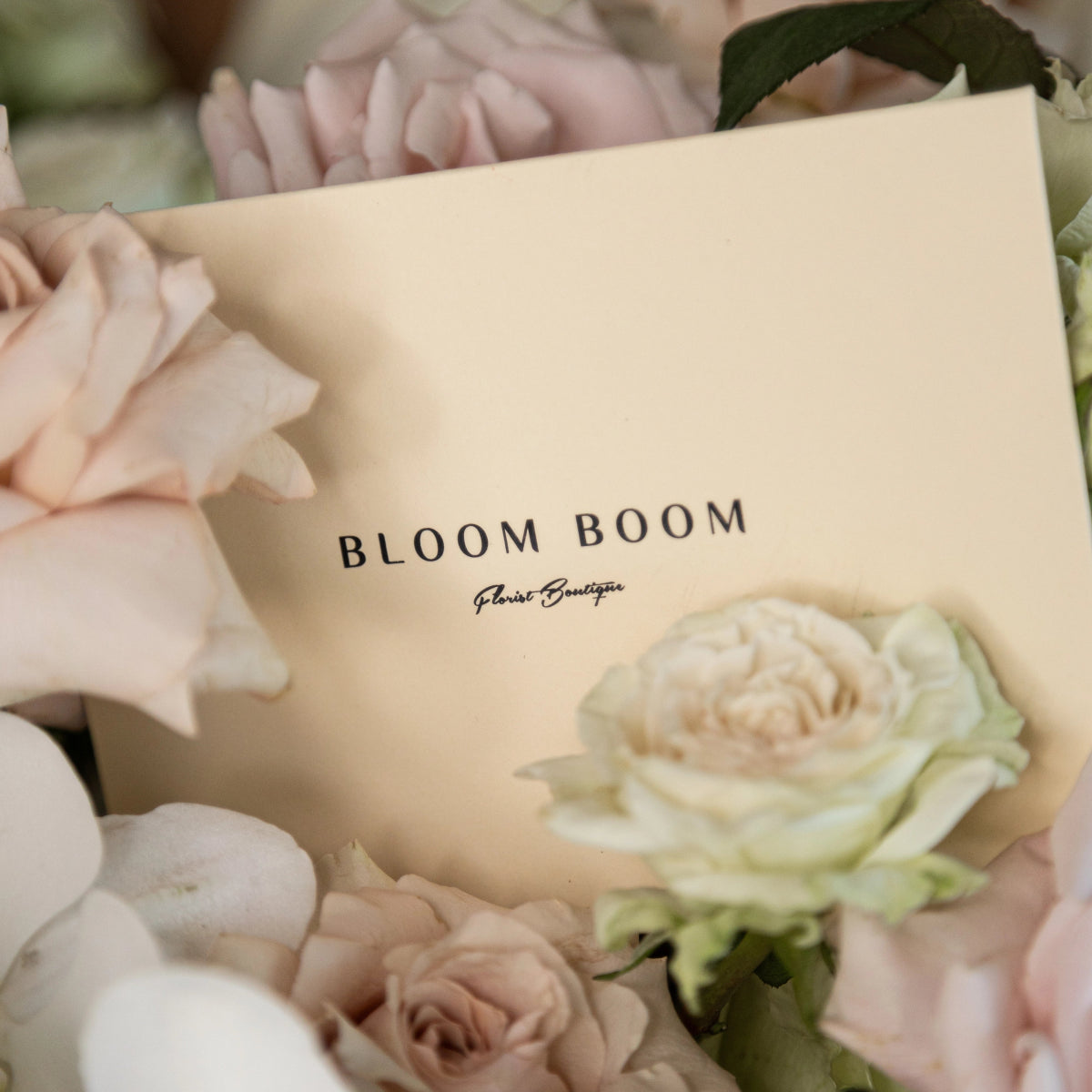 Cream colored Bloom Boom florist boutique gift box surrounded by soft pink roses and white ranunculus flowers