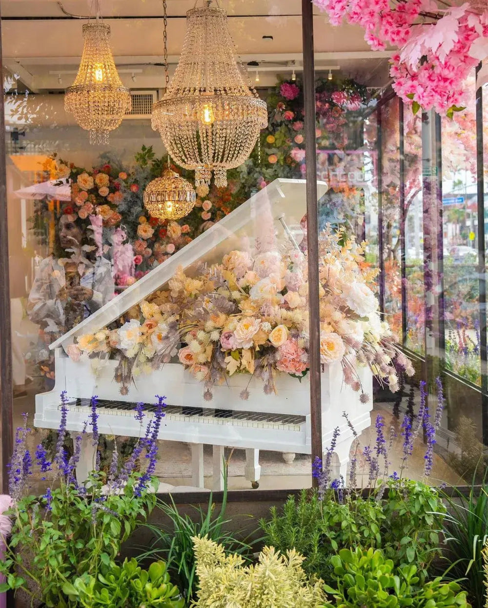 White grand piano dressed in blush florals framed by crystal chandeliers in the Bloom Boom West Hollywood storefront window