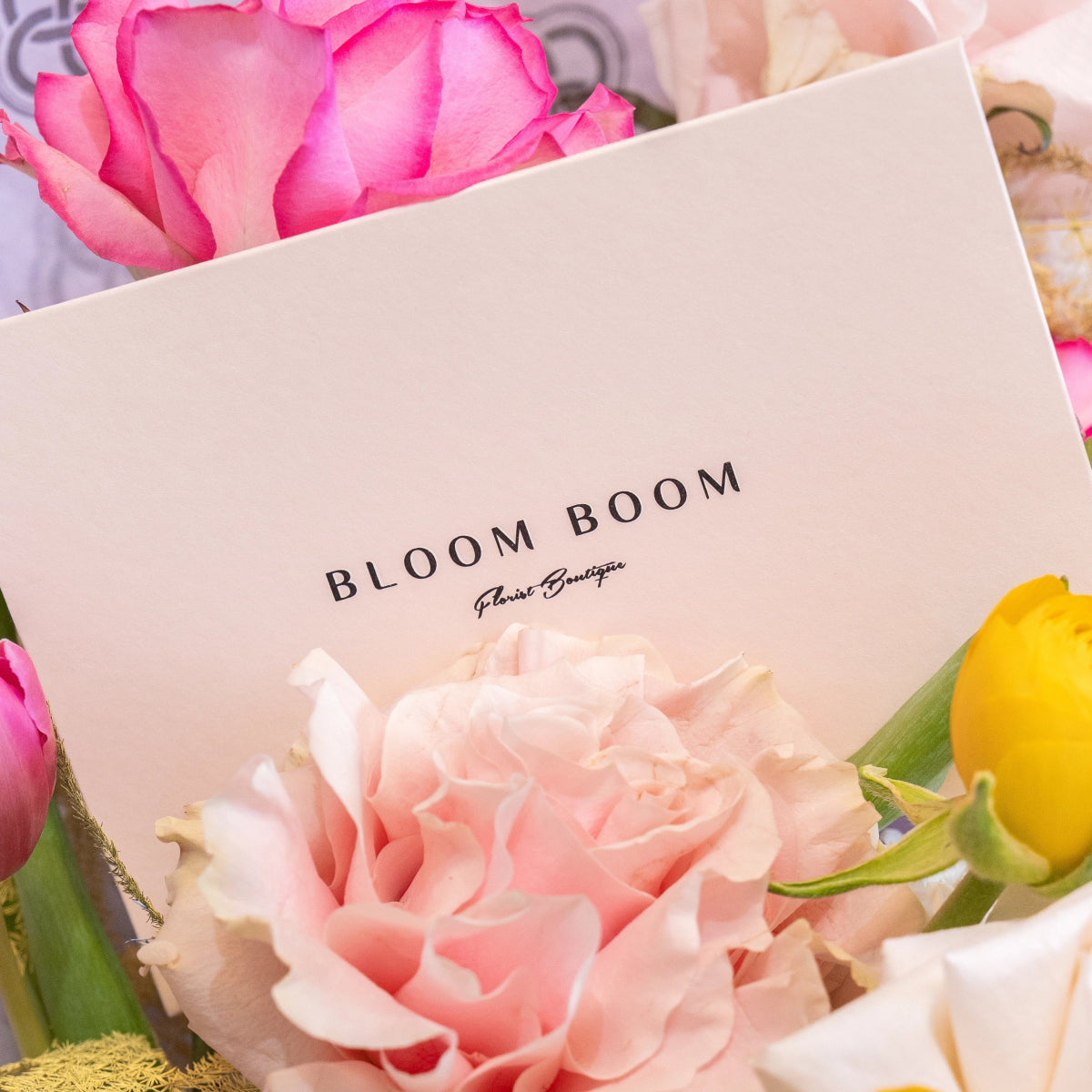 Bloom Boom Florist Boutique business card displayed with pink roses, peach carnations and yellow tulip arrangement