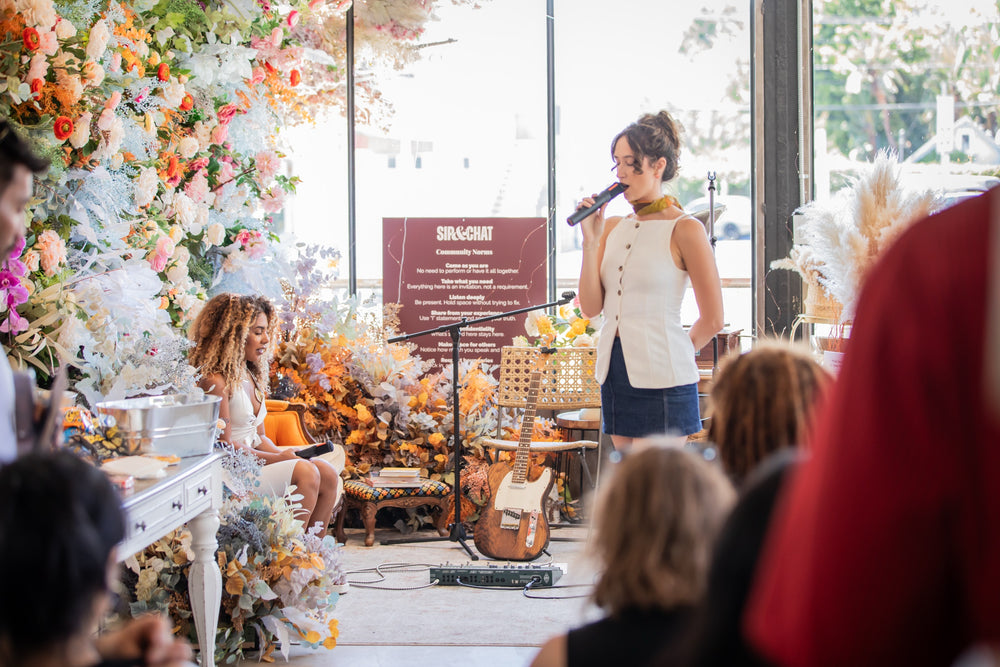 Performer at the microphone beside the white piano and floral installation, guests seated close