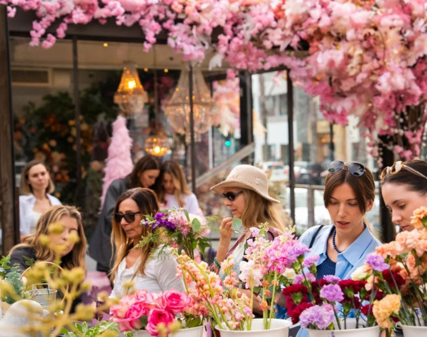 Women enjoying colorful flower arranging workshop under pink cherry blossoms at outdoor cafe terrace