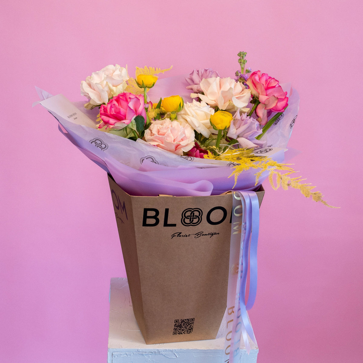 Colorful spring flower bouquet with pink roses, yellow tulips, and purple wrapping in brown BLOOM boutique bag
