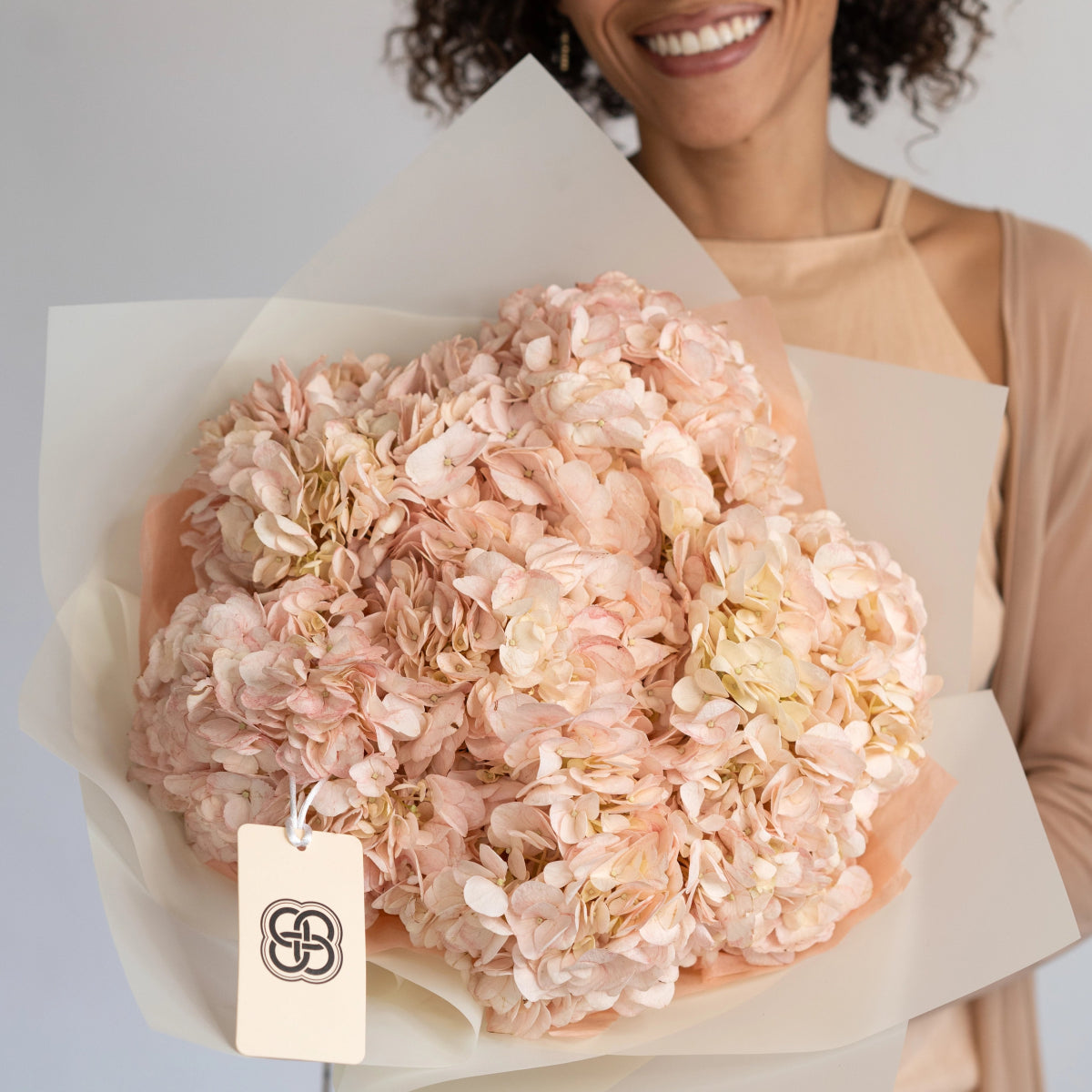 Blush pink and cream hydrangea bouquet wrapped in white paper held by smiling woman in peach dress