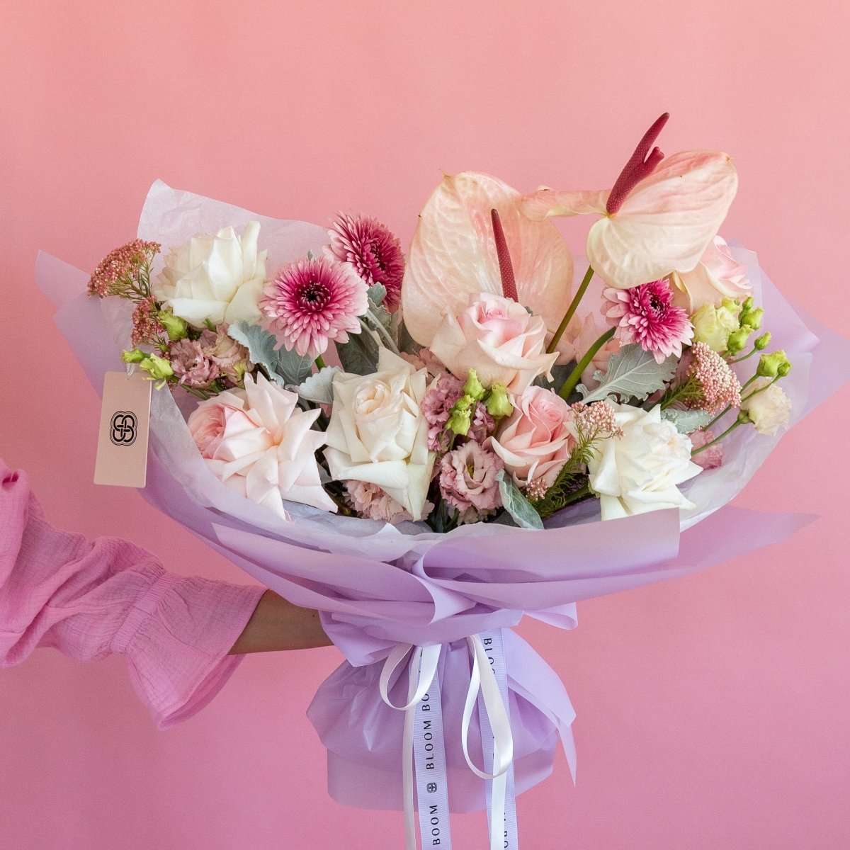 Pink and white flower bouquet with anthurium, roses, chrysanthemums wrapped in lavender paper on pink background