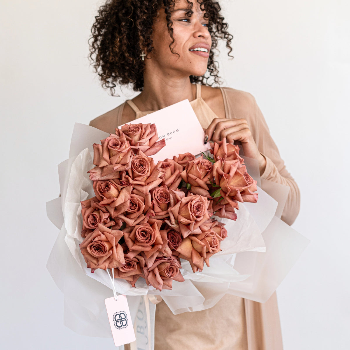 Large coral pink rose bouquet wrapped in white paper with woman in beige top holding flowers and pink gift card