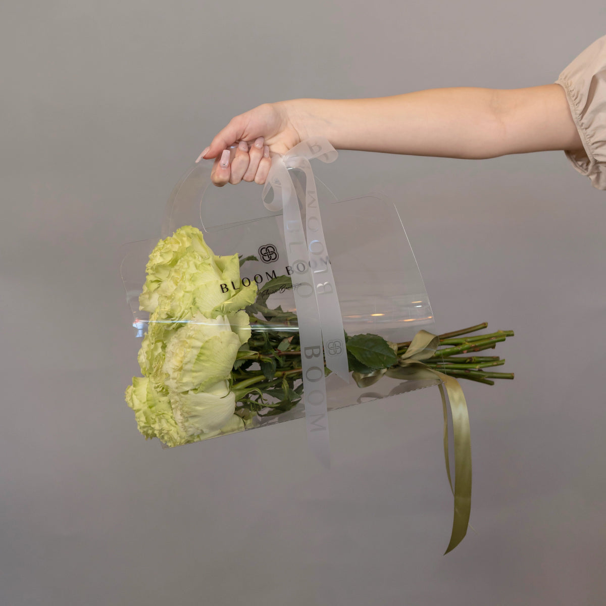 Hand holding clear tote bag with 12 green roses and gold ribbon against neutral background