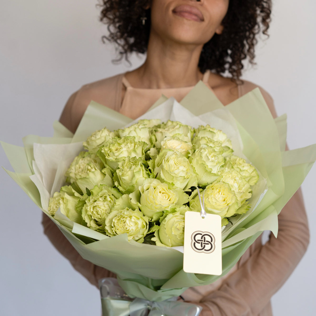 Light green roses bouquet wrapped in sage green paper with white tag, held by woman with curly hair