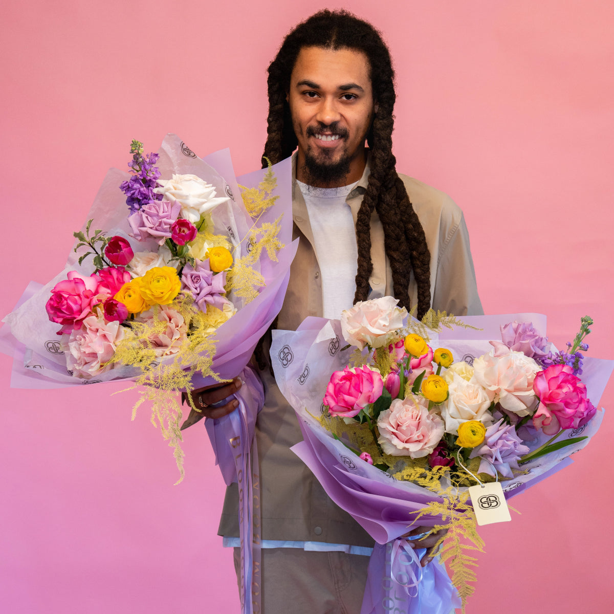 Smiling man holding two colorful mixed flower bouquets in purple wrapping with roses, ranunculus and yellow mimosa