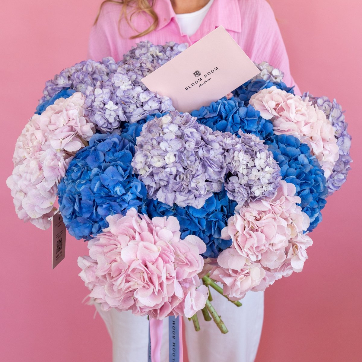 Mixed hydrangea bouquet with blue, purple and pink blooms held by woman in pink shirt with Bloom Boom card