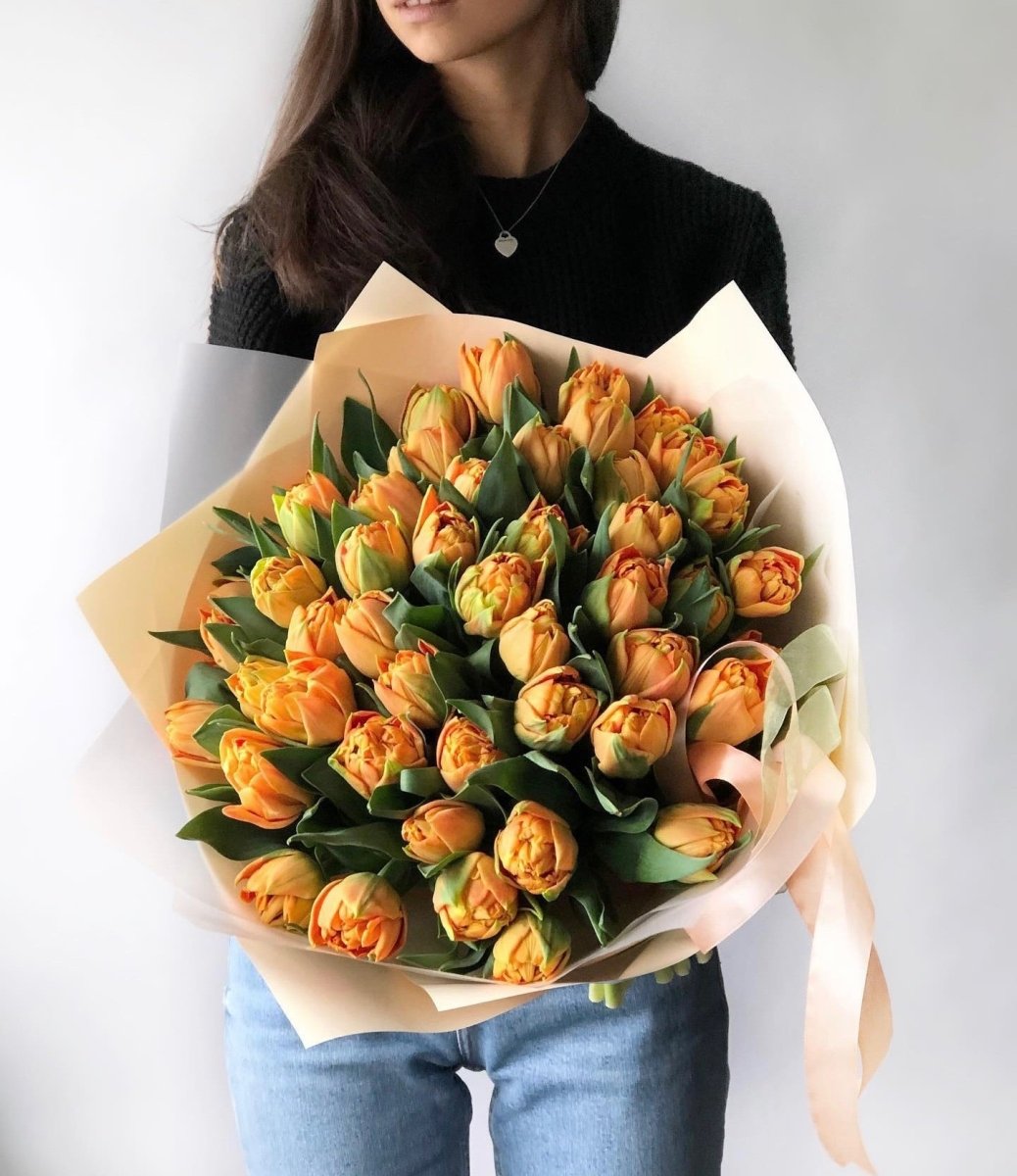 Large bouquet of 49 orange tulips wrapped in cream paper with ribbon, held by woman in black sweater