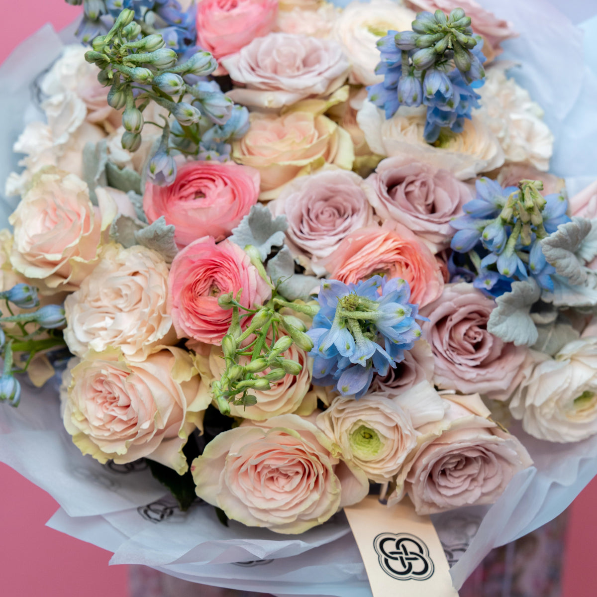 Pastel flower bouquet with pink roses, coral ranunculus, blue delphinium and dusty miller in white wrapping