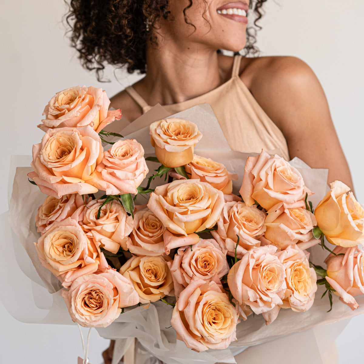 Smiling woman holding large bouquet of peach coral roses wrapped in white paper against neutral background