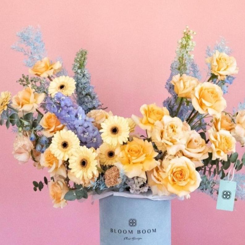 Peach roses and yellow gerberas with blue delphinium in light blue round flower box on pink background
