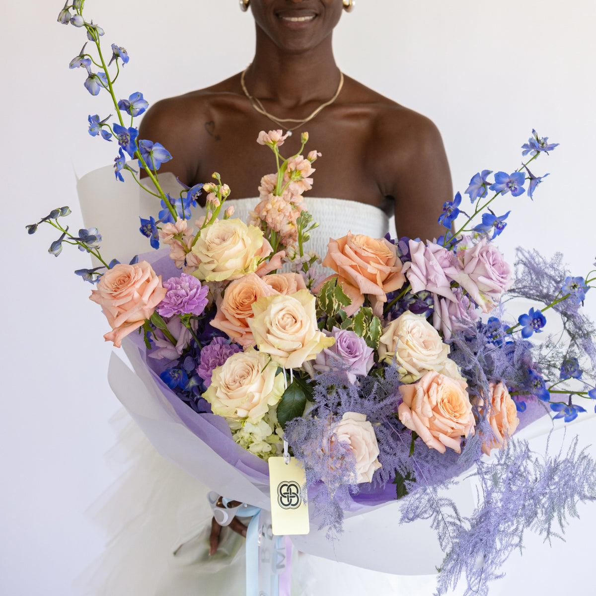 Mixed pastel bouquet with peach roses, purple blooms, blue delphiniums and dusty miller in lavender paper wrap