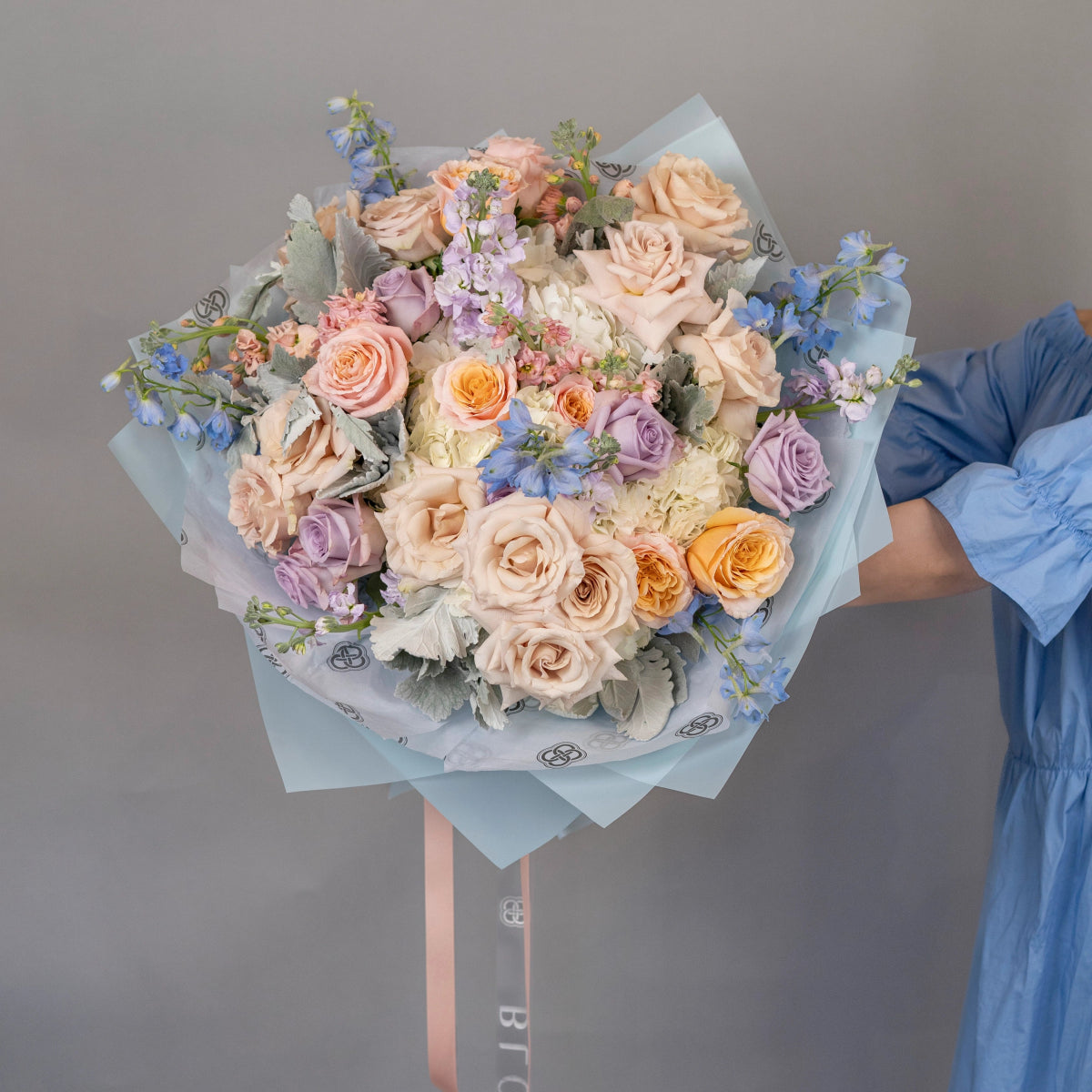 Mixed flower bouquet with peach roses, purple roses, blue delphinium, and eucalyptus in light blue wrapping paper