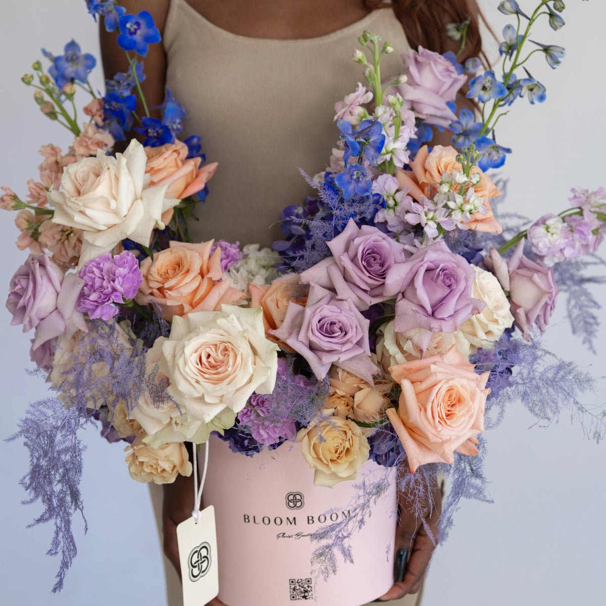 Pink round flower box with lavender roses, peach roses, blue delphiniums and dusty miller foliage arrangement