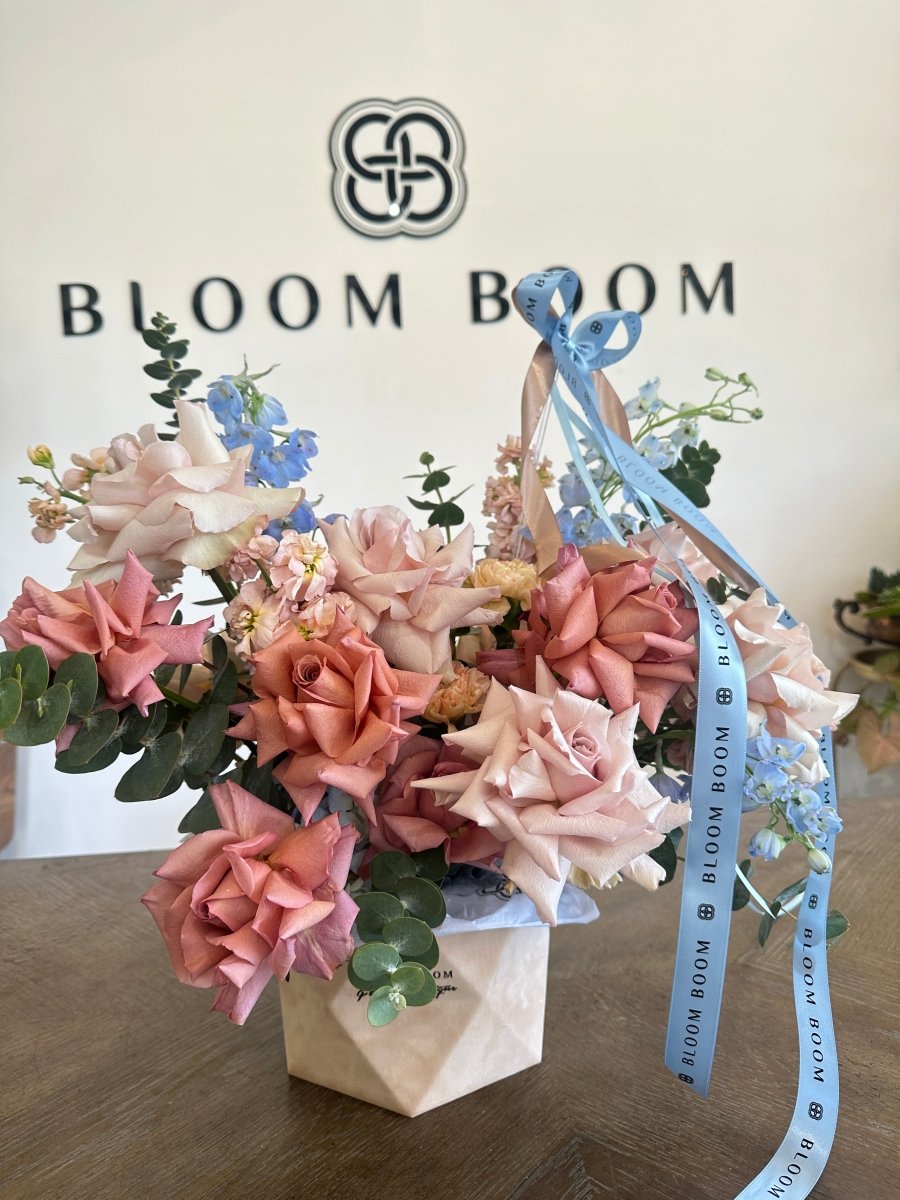 Pink and coral roses with blue delphinium in white geometric vase tied with Bloom Boom ribbon