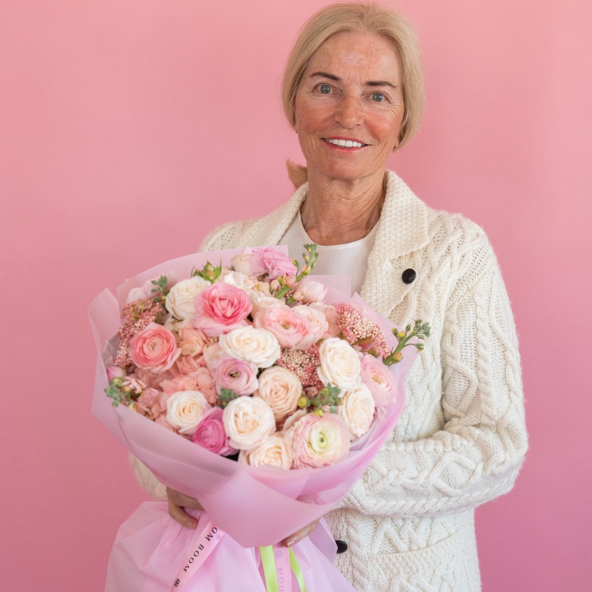 Pink and cream ranunculus bouquet in pink wrapping held by woman in cream cable knit cardigan