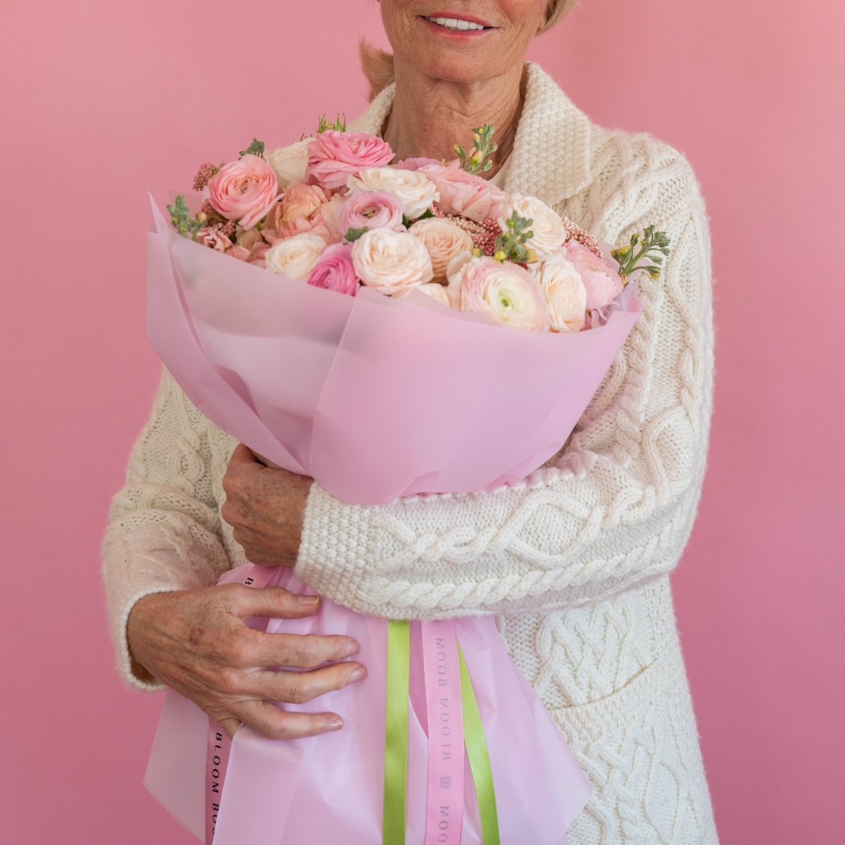 Pink ranunculus and rose bouquet in soft pink wrapping paper with green ribbon held by woman in cream knit sweater