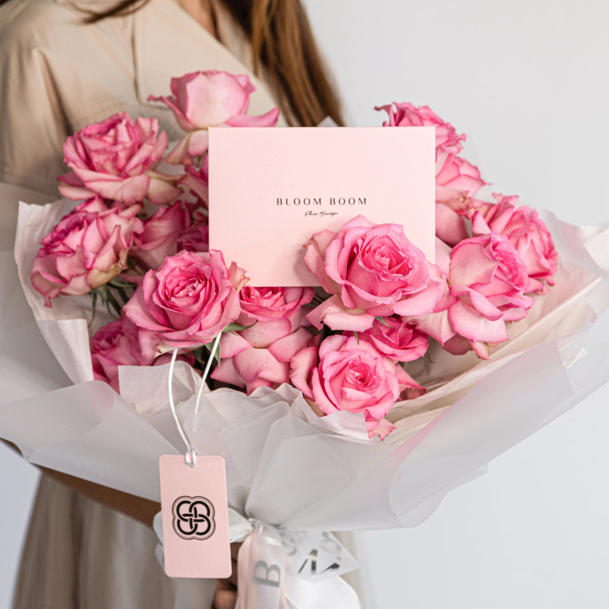 Pink rose bouquet wrapped in white paper with Bloom Boom branded card and gift tag held by woman in beige hoodie