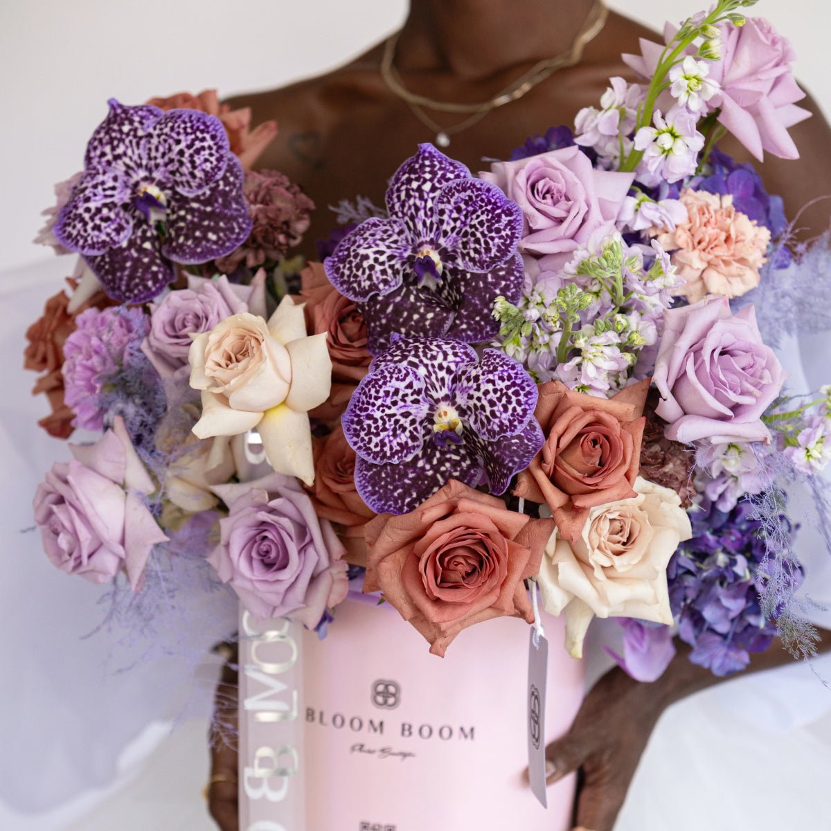 Purple spotted orchids with lavender roses, peach carnations in pink Bloom Boom gift box arrangement