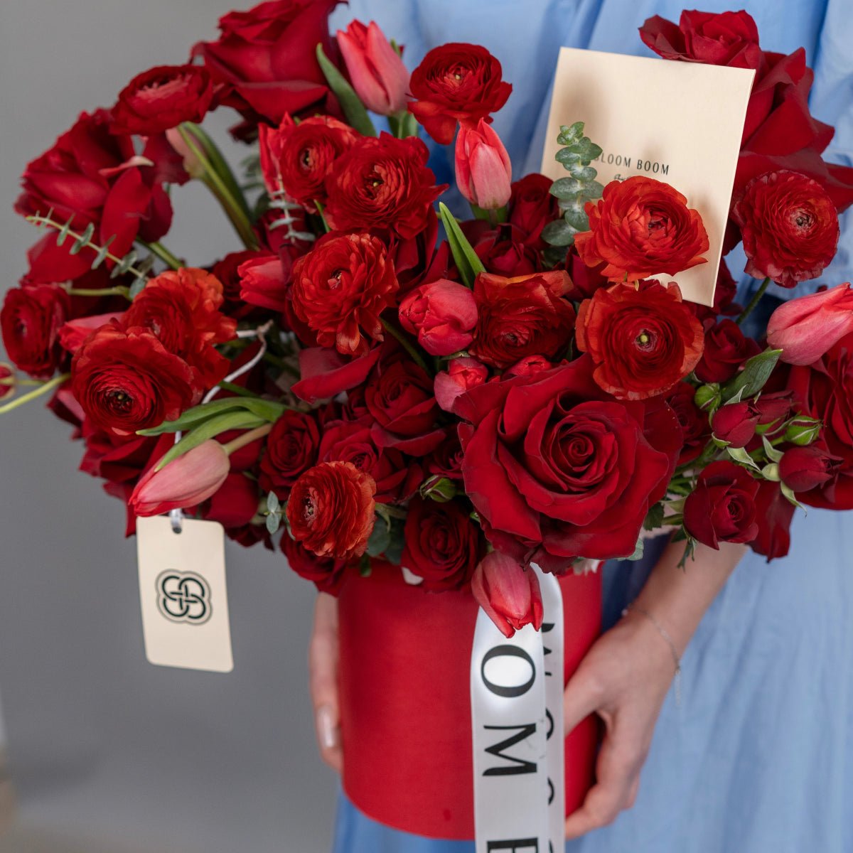 Red roses, ranunculus and pink tulips in white round flower box with ribbon and Bloom Boom card