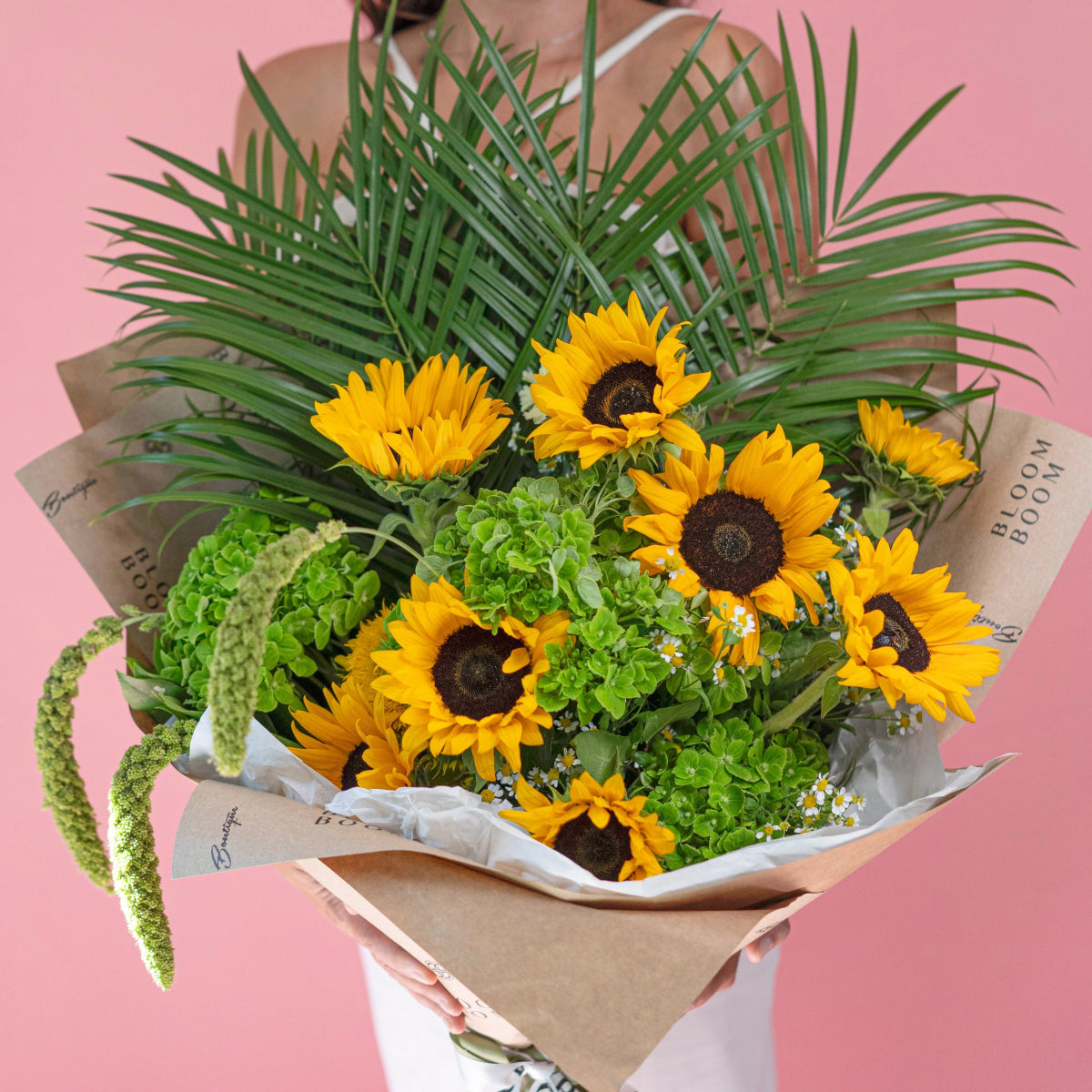 Bright yellow sunflower bouquet with green palm fronds and textured foliage wrapped in brown kraft paper