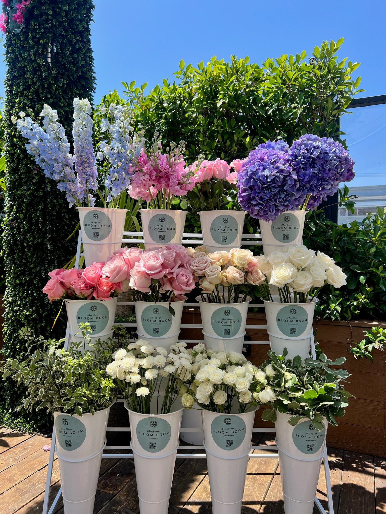Three-tier white metal flower display with purple hydrangeas, pink roses, white roses and blue delphiniums in buckets