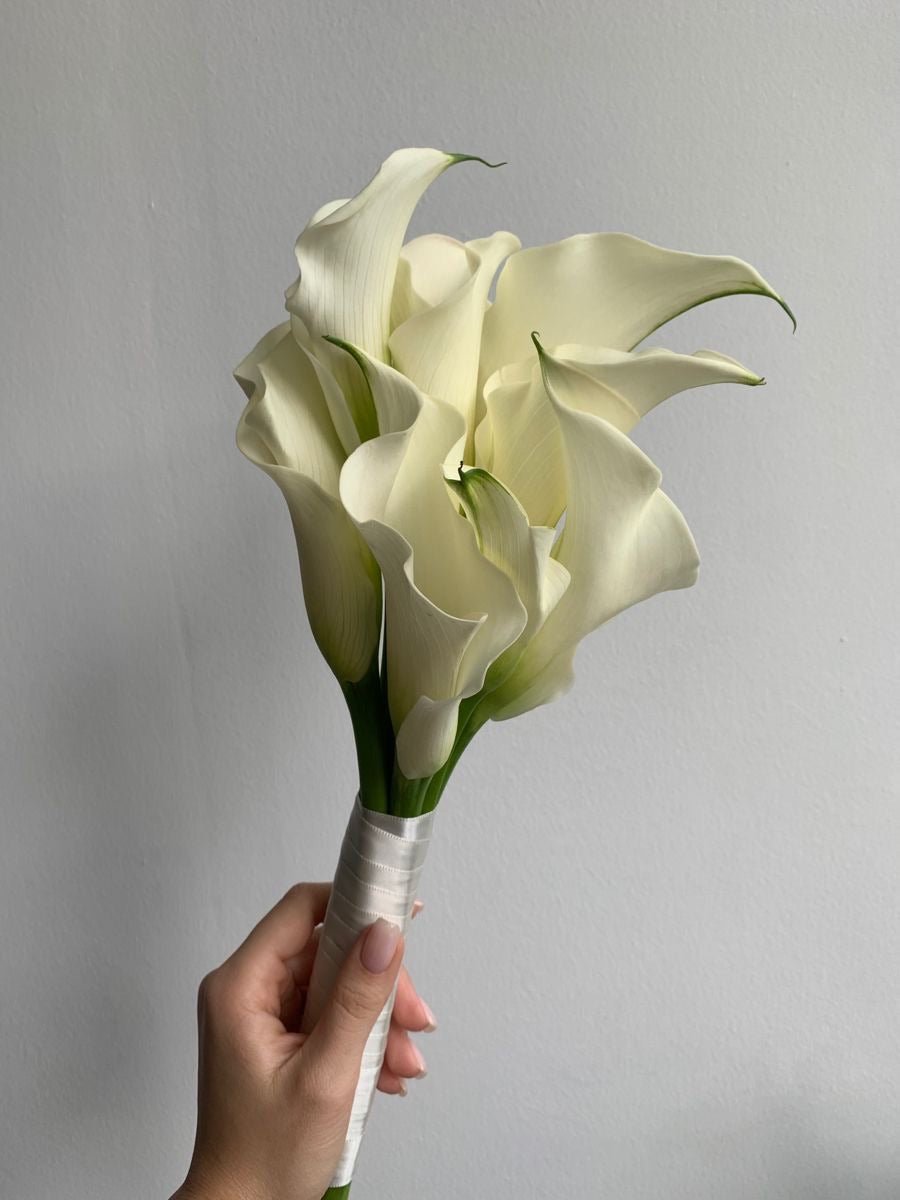 White calla lily bridal bouquet with satin ribbon wrap held in hand against gray background